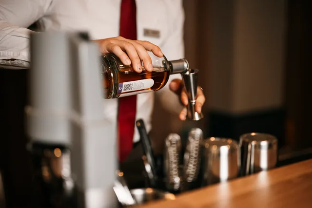 A bartender is pouring liquor from a bottle into a jigger behind the bar. The setting is a city hotel during the evening. The bar is well-equipped with various tools. Nonalcoholic bar equipment: four shifts, three to follow and one to skip